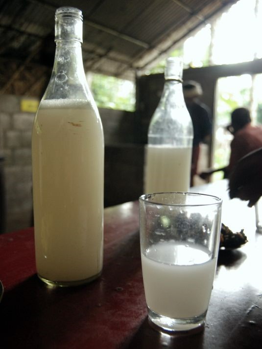 A bottle of palm wine sits on a table next to a glass of the same milky white substance.